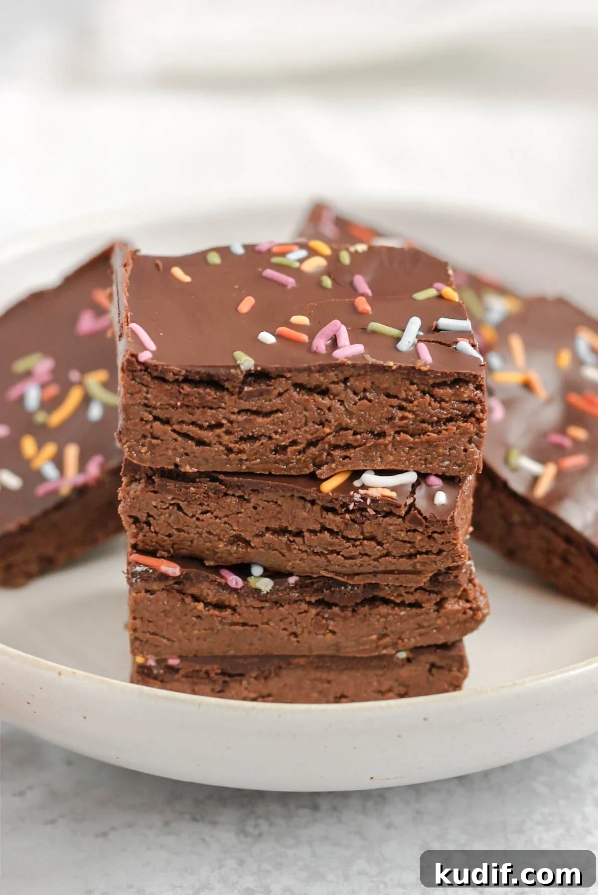 Square pieces of chocolate protein bar stacked 4 high on a plate with other bars in the background.