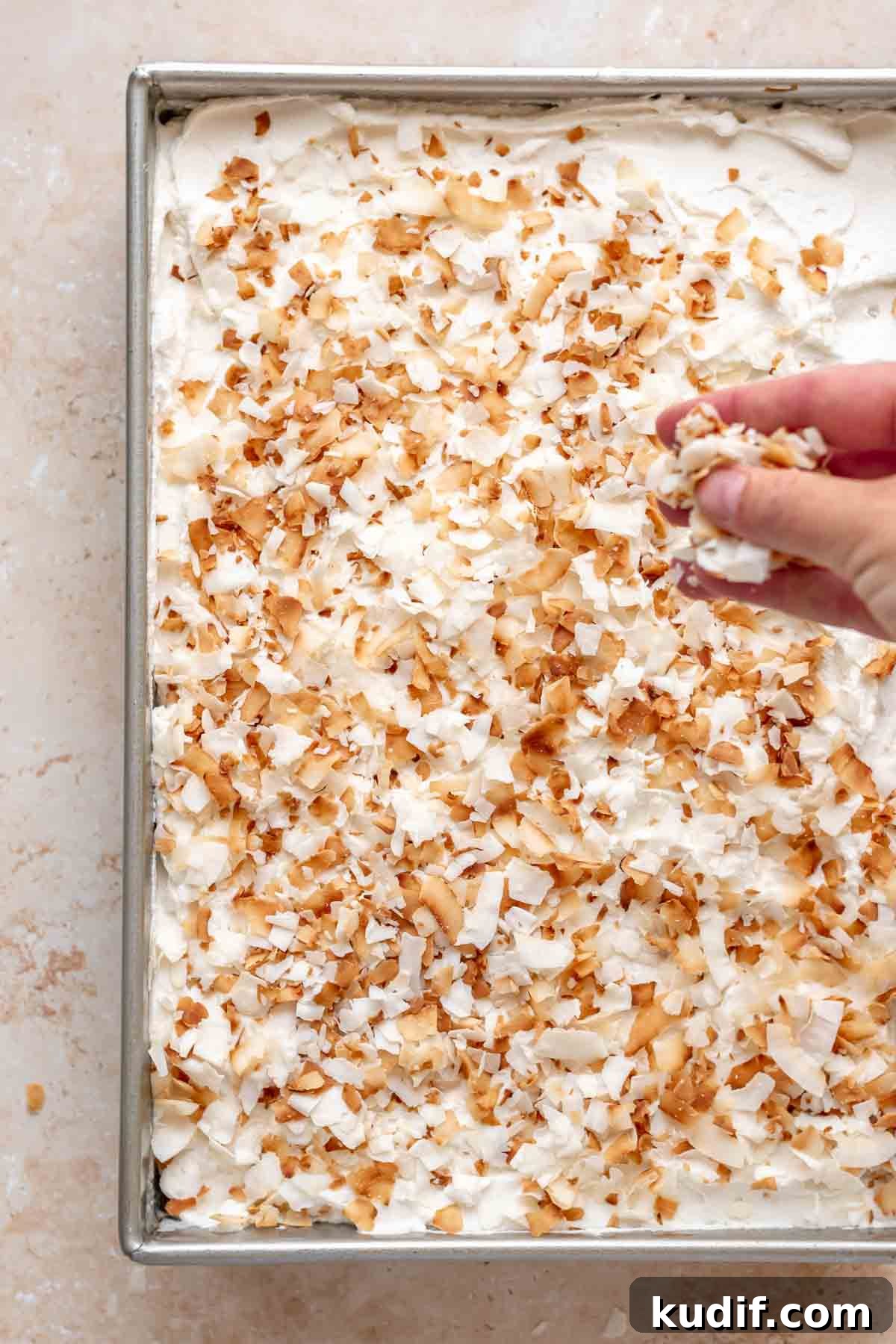Coconut Tres Leches Cake Recipe — Moist Latin-Style Dessert 16 A hand sprinkles toasted coconut on top of the whipped cream on the cake.