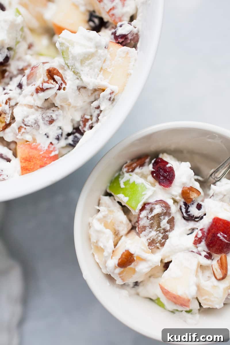 Creamy Holiday Fruit Salad with Cinnamon and Citrus 2 White bowls with creamy whipped cream topped fruit salad with red and green apples, grapes, bananas, cranberries and pecans.