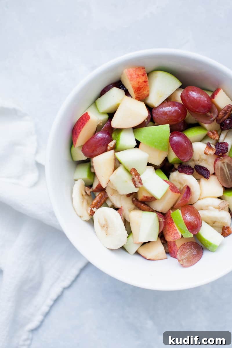 Creamy Holiday Fruit Salad with Cinnamon and Citrus 6 White bowl with red and green apples, pecans, grapes, bananas, and cranberries.