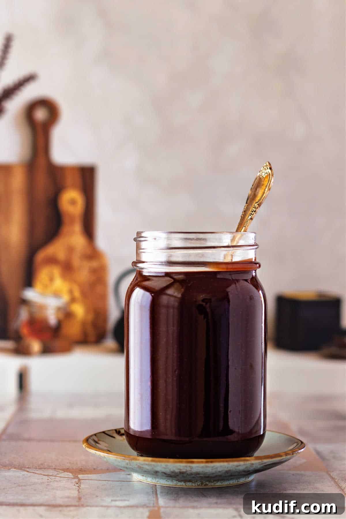 Classic Old-Fashioned Hot Fudge Sauce Recipe 2 A glass jar full of fudge sauce with a spoon in it. It's shot on small gray tiles with a couple of cutting boards propped up in the background.