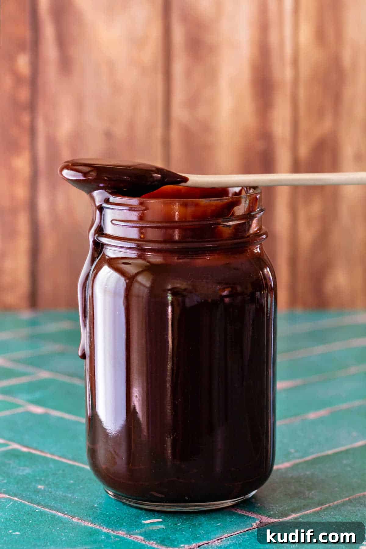 Classic Old-Fashioned Hot Fudge Sauce Recipe 3 A glass jar full of dark brown hot fudge sauce on a jade green tile surface with a beige wooden background. There is a white ceramic spoon balanced on top of the jar, and fudge sauce from the spoon is running down the side of the jar.