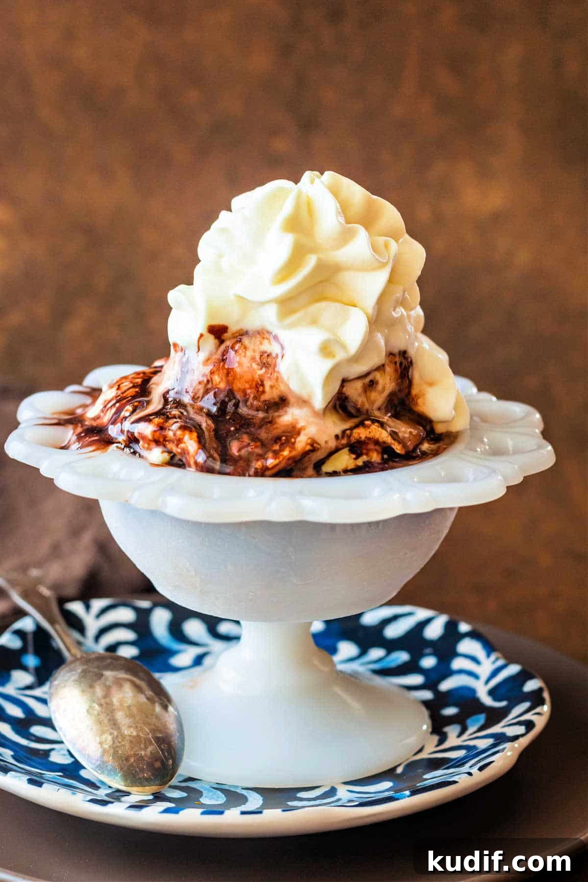 Classic Old-Fashioned Hot Fudge Sauce Recipe 9 A shot of ice cream in a white dish with chocolate syrup and whipped cream on top.