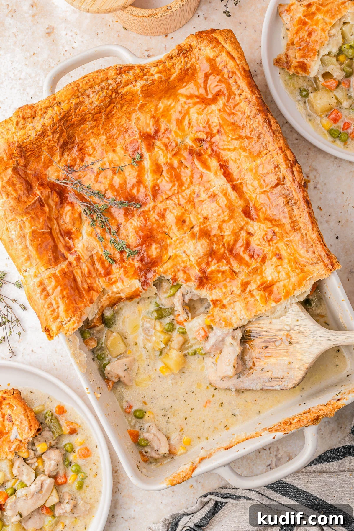 Comforting Chicken Pot Pie Casserole Recipe for Weeknights 2 Golden-brown puff pastry-topped chicken pot pie in a baking dish, partially served with filling of chicken and mixed vegetables, surrounded by two plated servings.