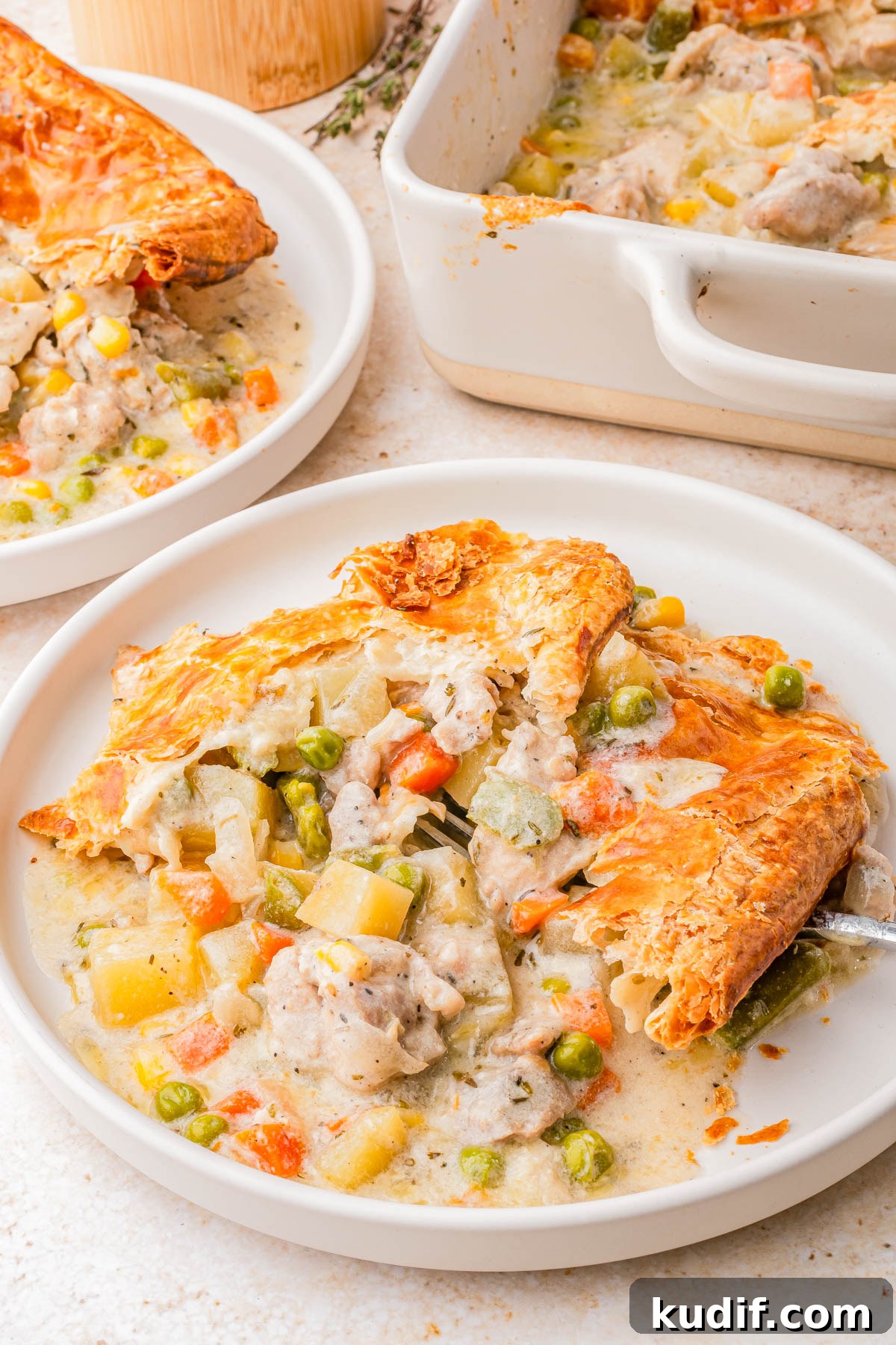 Comforting Chicken Pot Pie Casserole Recipe for Weeknights 4 A serving of chicken pot pie with a golden, flaky crust on a white plate, showing mixed vegetables and creamy filling; a baking dish and another plate are in the background.