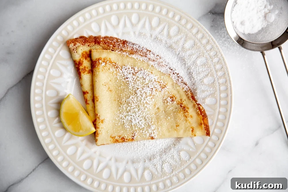 Crepes on a plate dusted with powdered sugar, with a lemon wedge alongside.