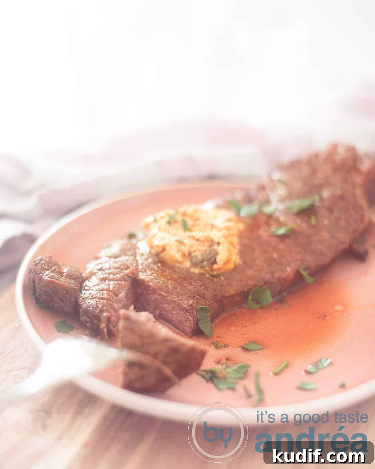 Crispy Air Fryer Sirloin Steak in 15 Minutes 6 Een hoogtefoto met een roze bord op een houten plank. Een lapje entrecote met een stukje er van af gesneden met een vork. Kruidenboter smelt erbovenop