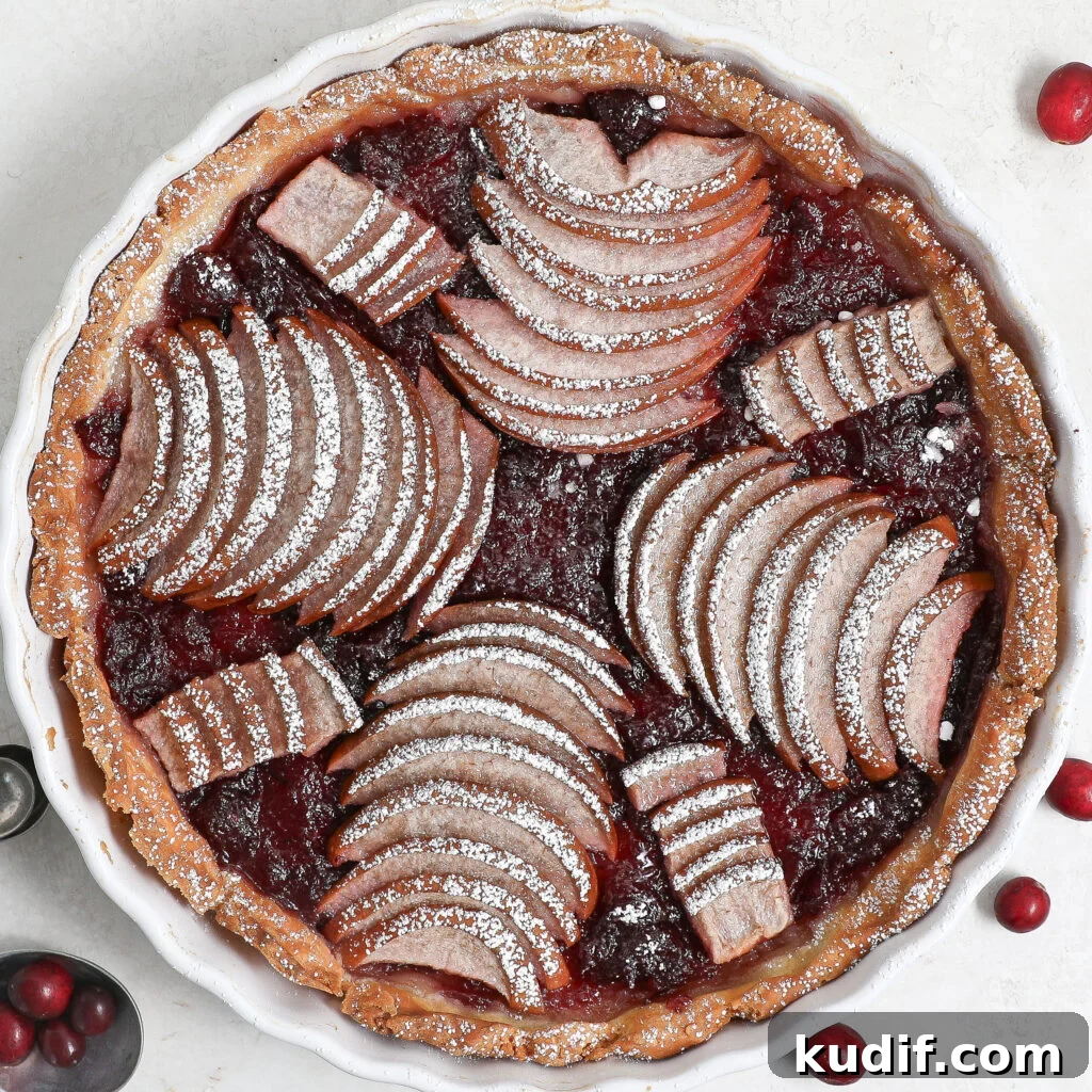An overhead view of a Pear Cranberry Tart with arranged slices of pears and cranberries, dusted with powdered sugar, in a round white dish on a light surface. Some whole cranberries are scattered nearby.