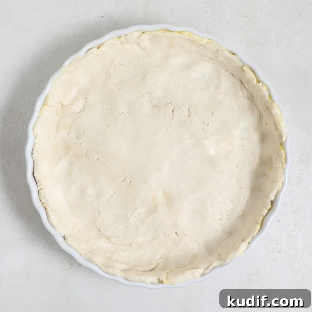 An unbaked pie crust for a Pear Cranberry Tart is evenly pressed into a round white ceramic pie dish, with crimped edges, sitting on a light-colored surface.