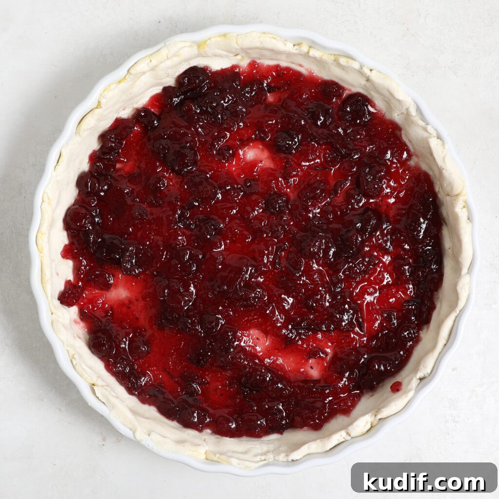 A round pie dish filled with unbaked crust and topped with a layer of vibrant red pear cranberry tart filling, seen from above on a white surface.