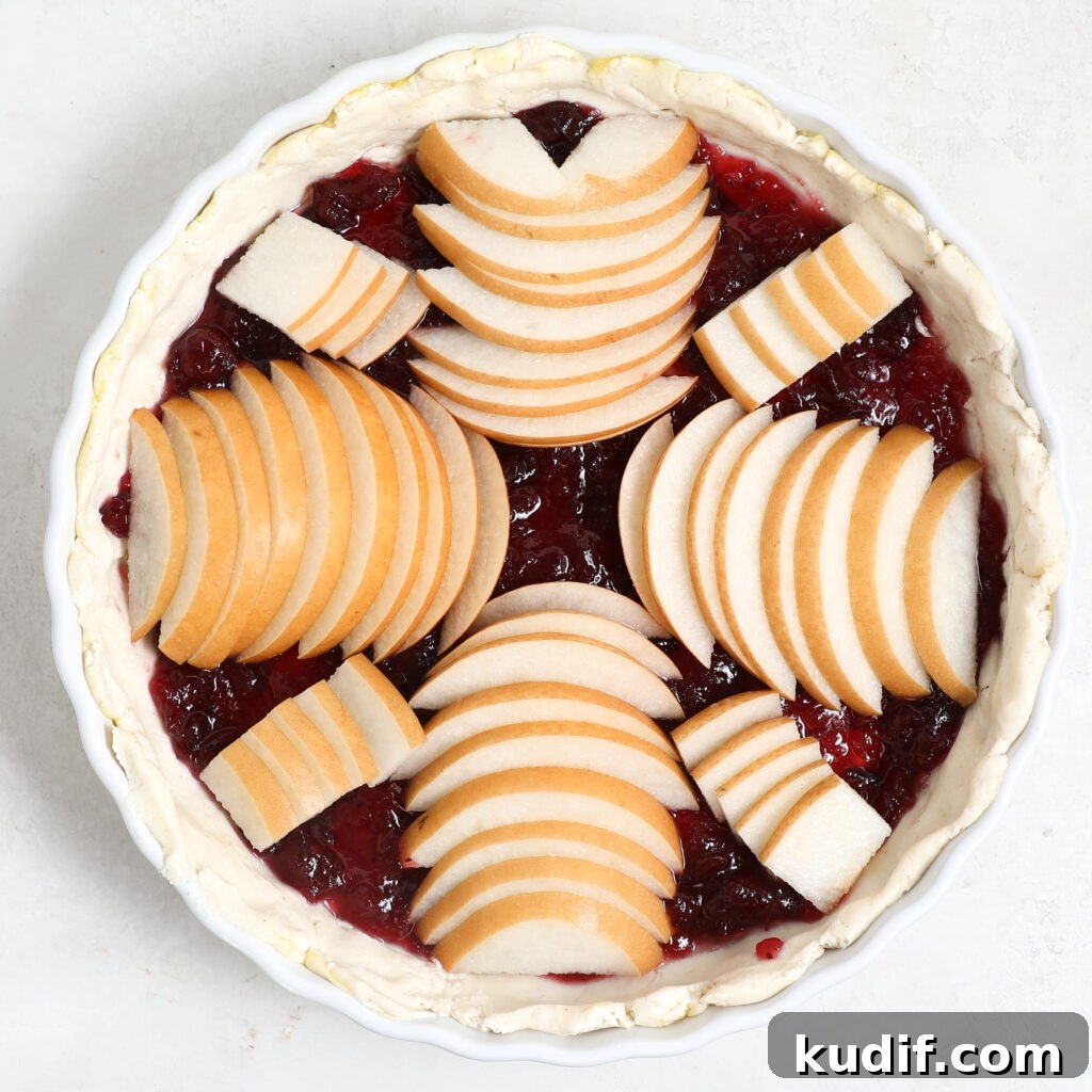 A Pear Cranberry Tart with a flaky crust, filled with dark red fruit and topped with fan-shaped pear slices arranged in a circular pattern before baking.