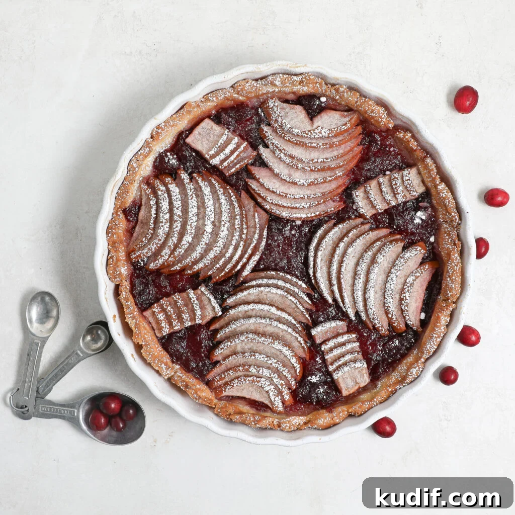 A Pear Cranberry Tart topped with fanned apple slices and powdered sugar sits in a white dish, while cranberries and metal measuring spoons rest nearby on a light surface.