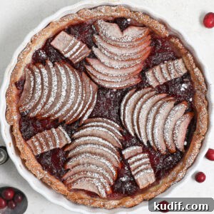 An overhead view of a Pear Cranberry Tart with arranged slices of pears and cranberries, dusted with powdered sugar, in a round white dish on a light surface. Some whole cranberries are scattered nearby.