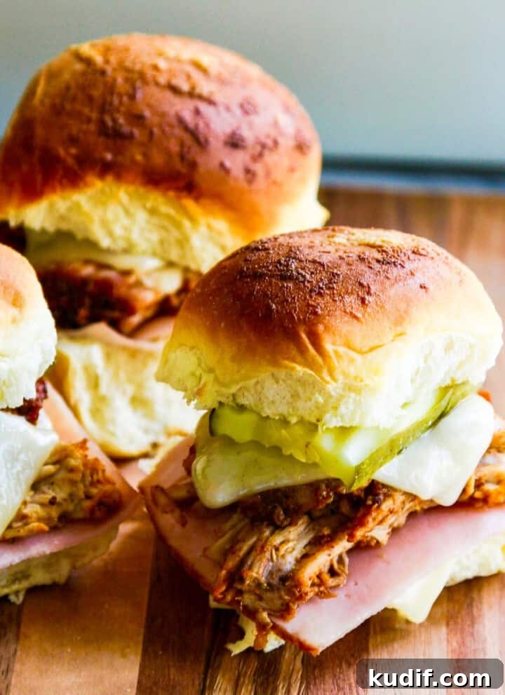 Three pulled pork sandwiches on Hawaiian buns with pickles, cheese and ham on a cutting board.