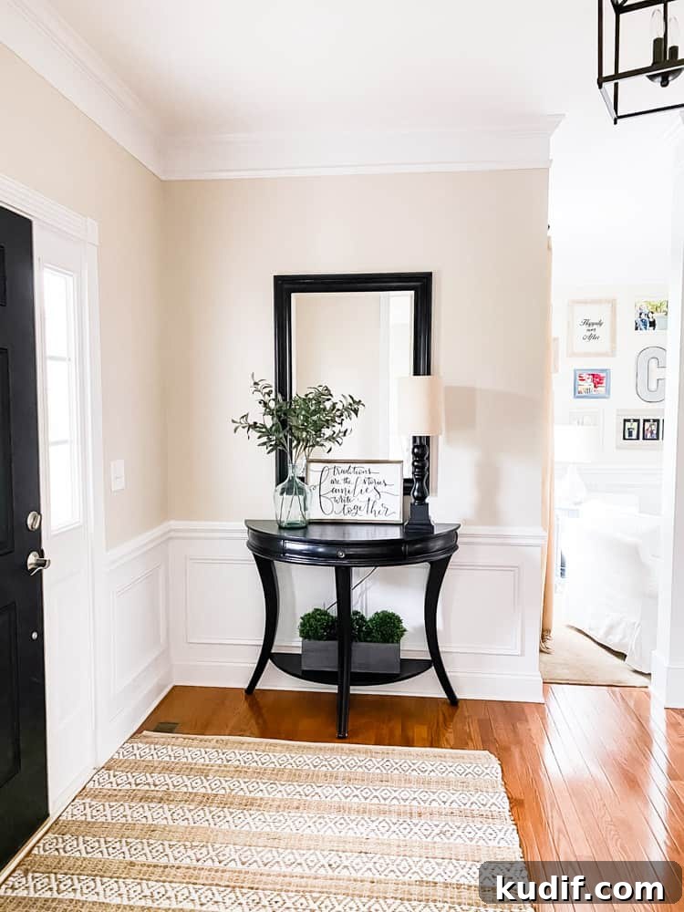 demi round table against wall in entryway next to front door with rectangular mirror hung above table. faux stems and plants decorate table and shelf on bottom of table. lamp also on table
