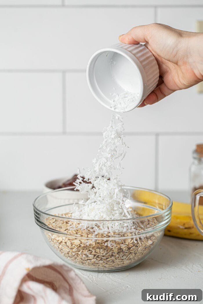 Dumping shredded coconut into a glass mixing bowl of oats. 