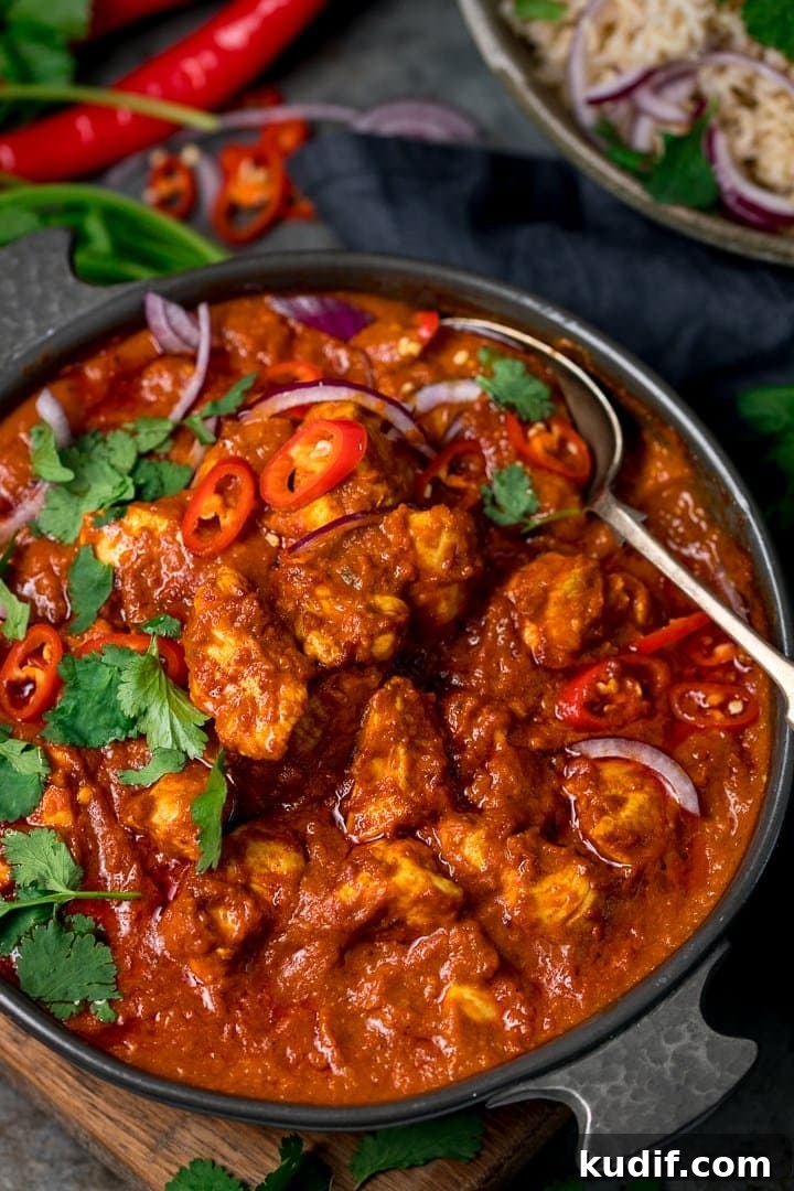 Authentic Chicken Madras Curry Recipe for Bold Flavor 8 chicken madras in a dark serving bowl with a spoon in the bowl. The madras is topped with coriander and sliced red onion and red chillies