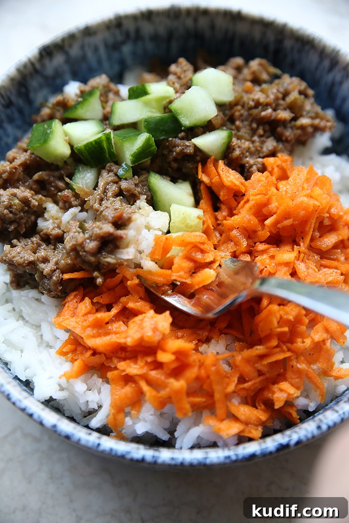 A bowl of rice with chopped cucumbers on top of ground beef and next to carrot kimchi. A spoon is inserted diagonally from the right into the carrot kimchi.