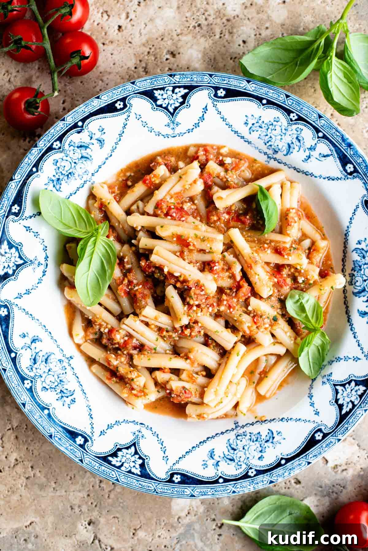 Fresh No-Cook Tomato Pasta Sauce Recipe for Quick Dinners 2 A blue and white vintage bowl with pasta in a fresh tomato sauce topped with basil. The bowl is on a travertine surface with cherry tomatoes and basil around.