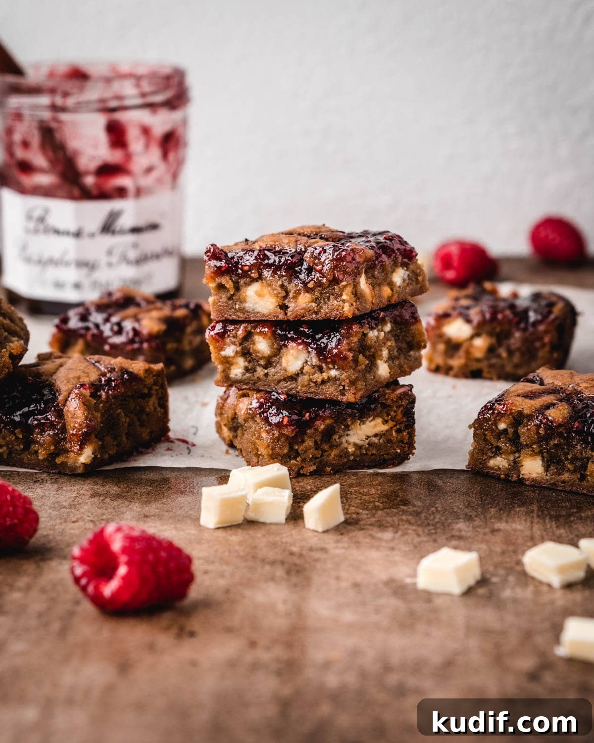 White Chocolate Raspberry Blondies with Tangy Fruit Swirls 13 Whole raspberries and white chocolate chunks laying in front of squares of white chocolate and raspberry blondies.