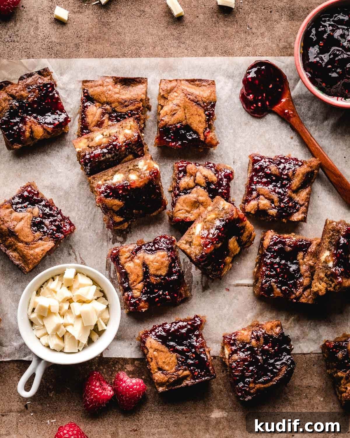 White Chocolate Raspberry Blondies with Tangy Fruit Swirls 3 Top view of white chocolate and raspberry blondies on parchment paper with small bowls of white chocolate chips and raspberry sauce next to them.