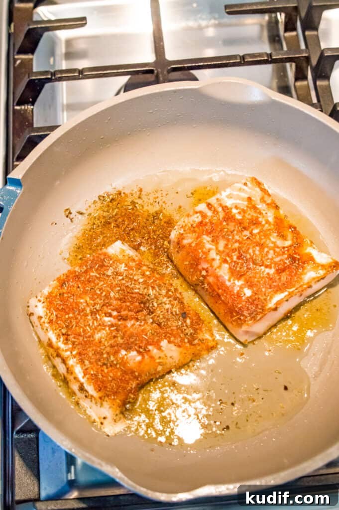 Two seasoned cod fillets cooking in a pan on the stovetop.