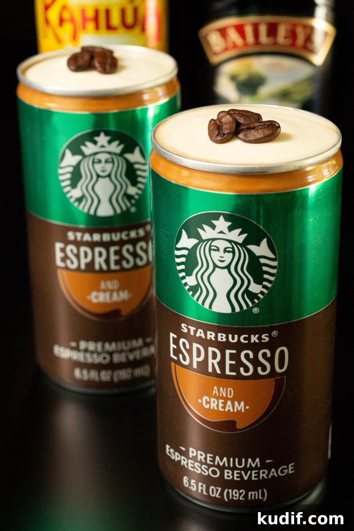 In focus in the foreground is a Starbucks Espresso and cream can. The top of the can has been removed to show a cold foam topping with three espresso beans sitting on top. Another can is out of focus in the background along with a bottle of Kahlua and Bailey's.