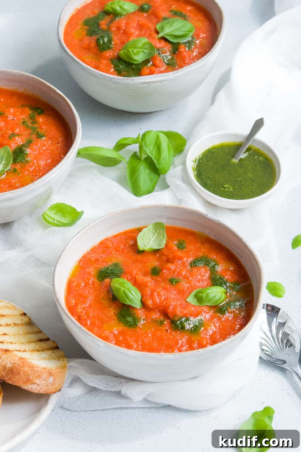 Roasted Red Pepper Soup Recipe — Creamy Roasted Capsicum Soup 2 three bowls of soup, some toasted bread on the side, and a dish of basil oil on the edge with some spoons and basil leaves scattered.