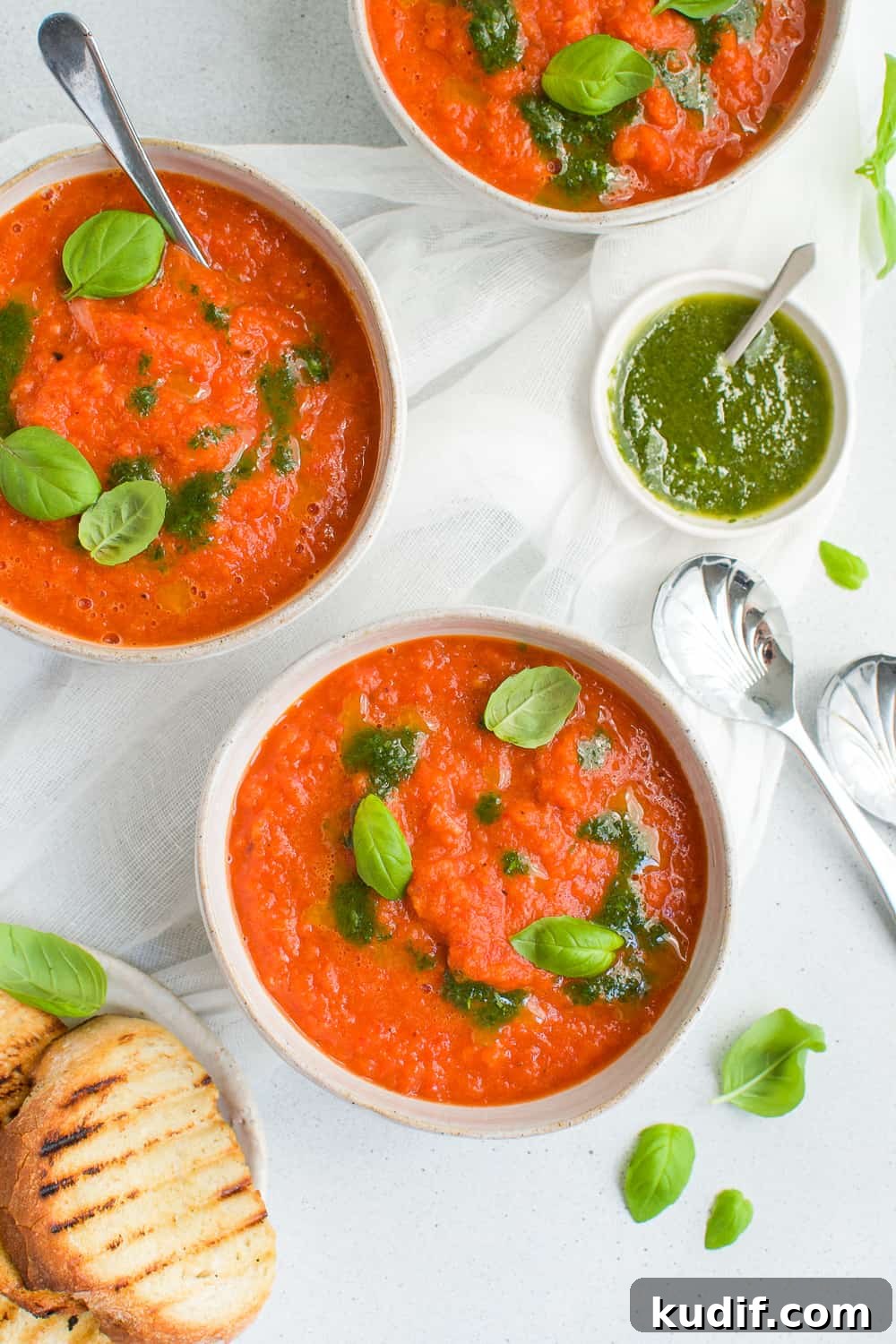 Roasted Red Pepper Soup Recipe — Creamy Roasted Capsicum Soup 5 three bowls of soup, some toasted bread on the side, and a dish of basil oil on the edge with some spoons and basil leaves scattered.
