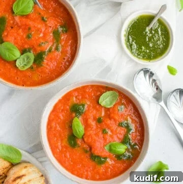 three bowls of soup, some toasted bread on the side, and a dish of basil oil on the edge with some spoons and basil leaves scattered.