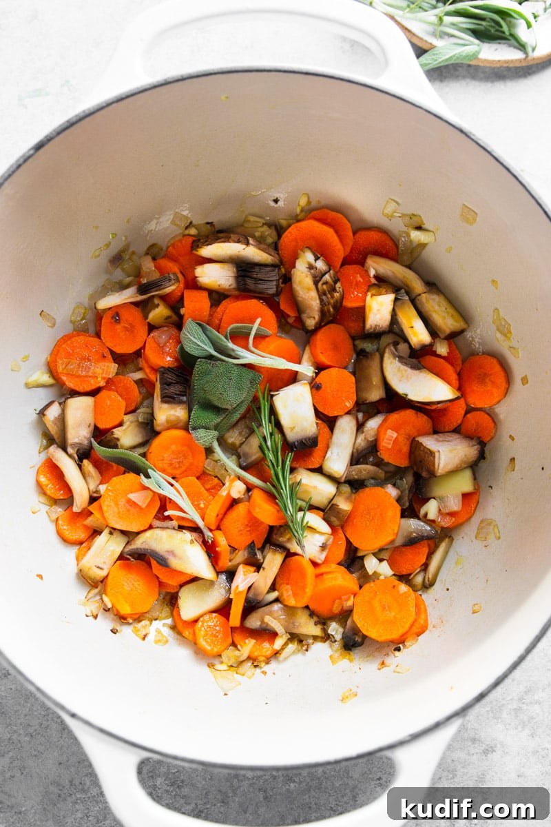 Hearty Turkey, White Bean, and Kale Soup Recipe 6 Carrots, mushrooms, onion, garlic and fresh rosemary and sage in a white pot.