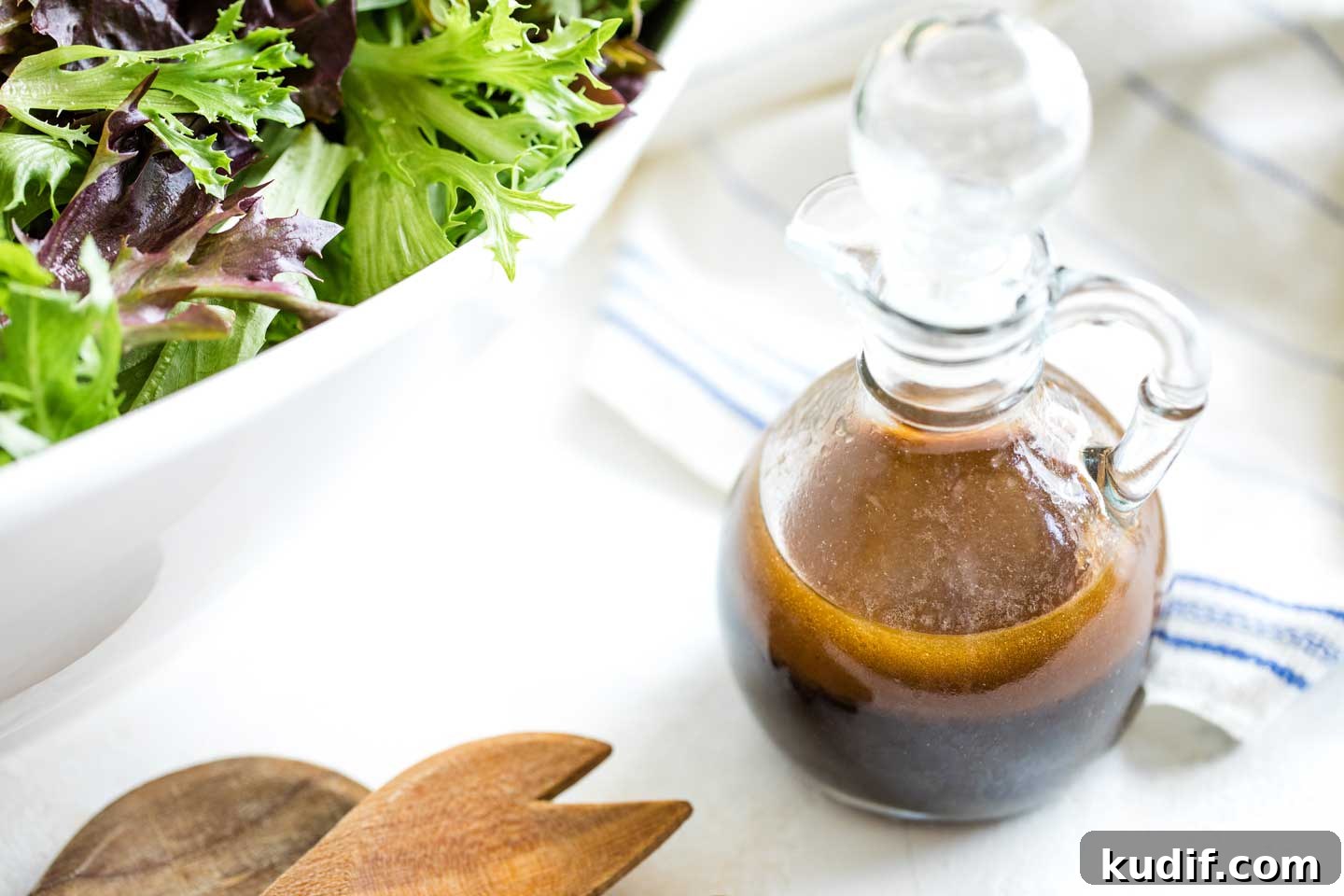 Healthy Balsamic Vinaigrette Recipe with Maple Syrup glass cruet of balsamic dressing with the stopper placed in the top, sitting next to wooden salad servers and a big bowl of salad