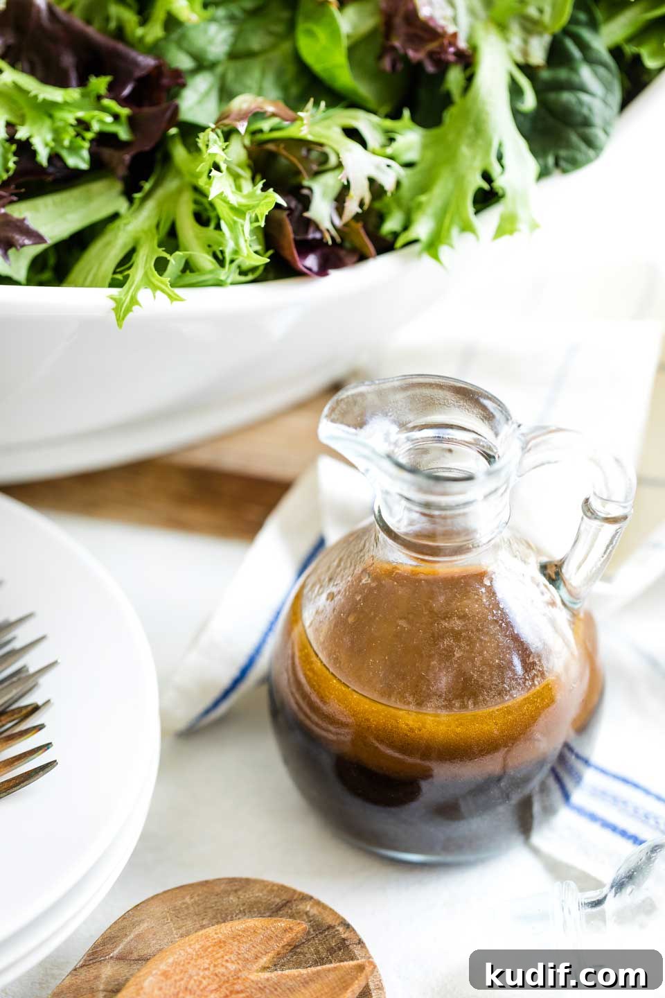 Maple Balsamic Vinaigrette Recipe cruet of dressing nestled near tea towel with navy stripe, with large bowl of salad, stack of plates and salad forks nearby