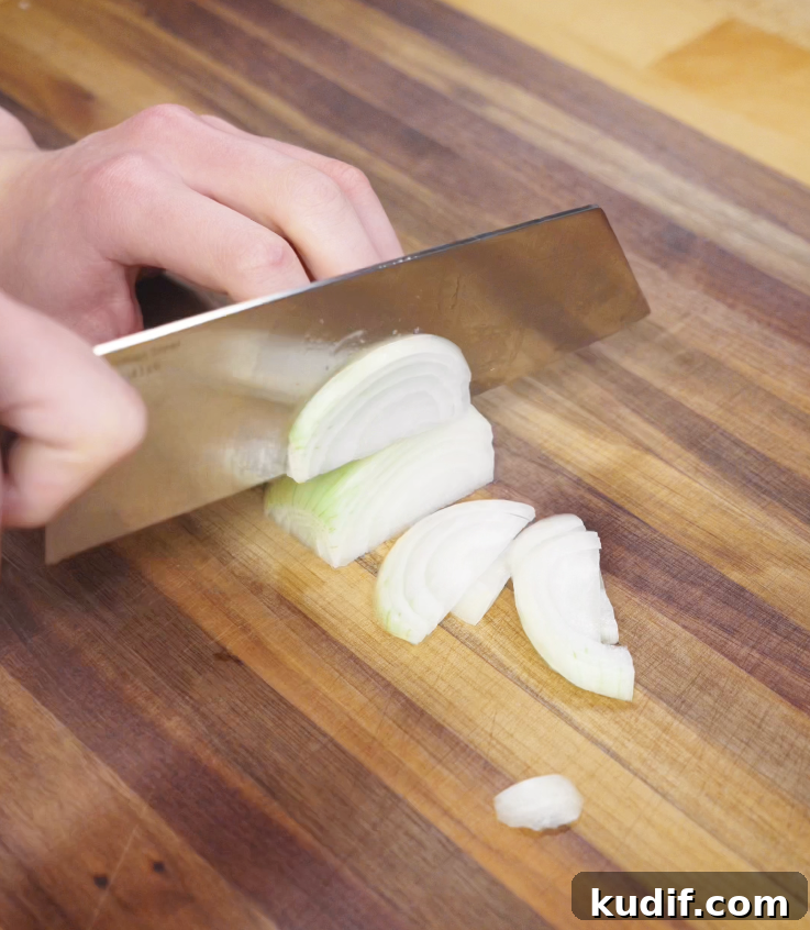 Authentic Chicken Chow Mein Recipe: Quick Stir-Fry Noodles 4 A hand slices a white onion into thin strips on a wooden cutting board using a large chef’s knife.