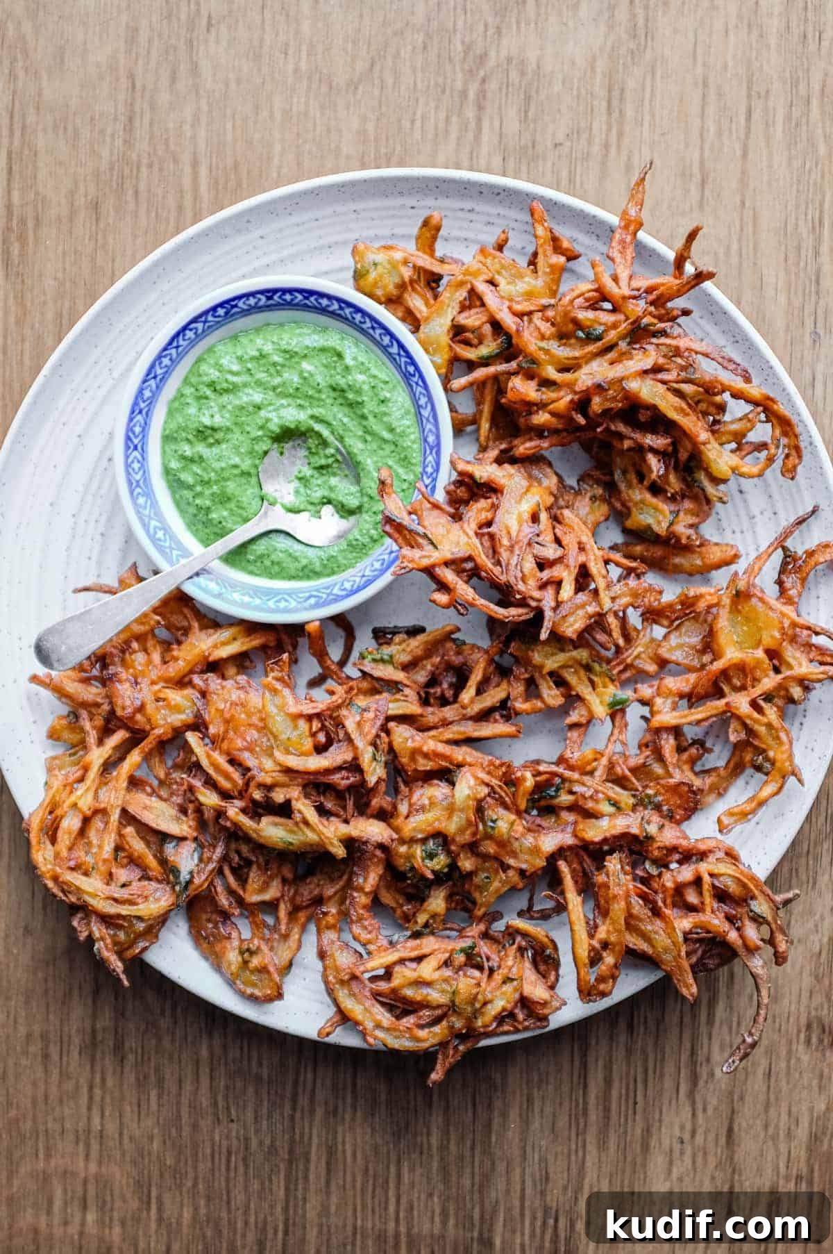 Onion Pakoda | cookeatworld.com Fried onion pakodas on a platter with a small bowl of coriander chutney and a small spoon.