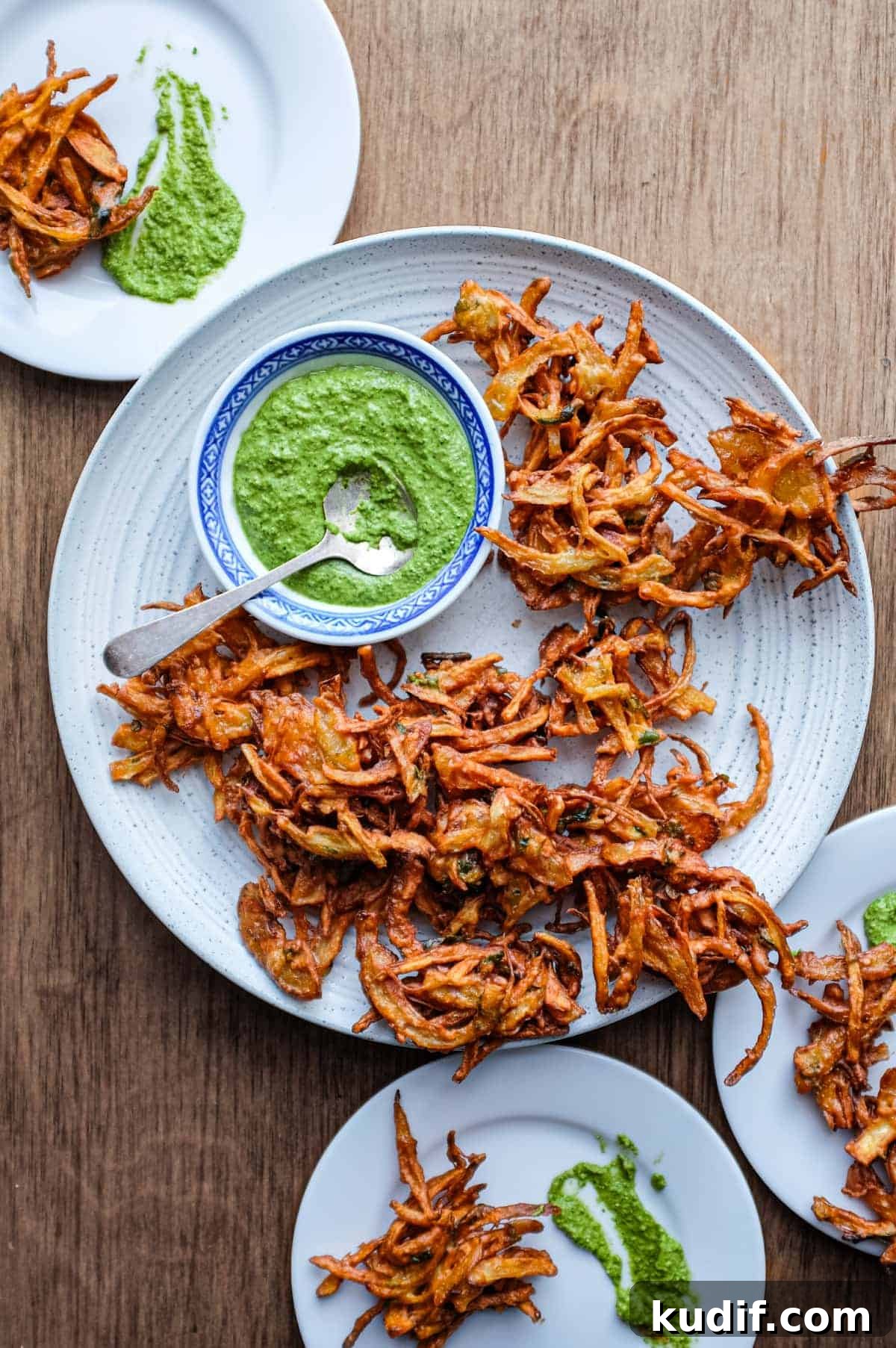Crispy Onion Pakoda Recipe: Indian Onion Fritters Guide 8 Fried onion pakodas on a platter with a small bowl of coriander chutney and a small spoon.