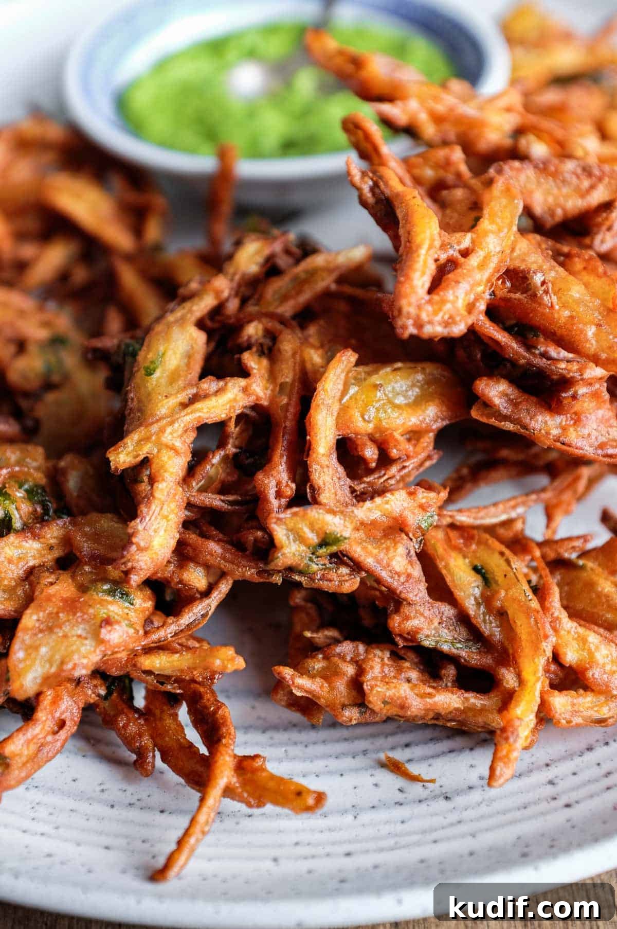 Crispy Onion Pakoda Recipe: Indian Onion Fritters Guide 9 Fried onion pakodas on a platter with a small bowl of coriander chutney and a small spoon.