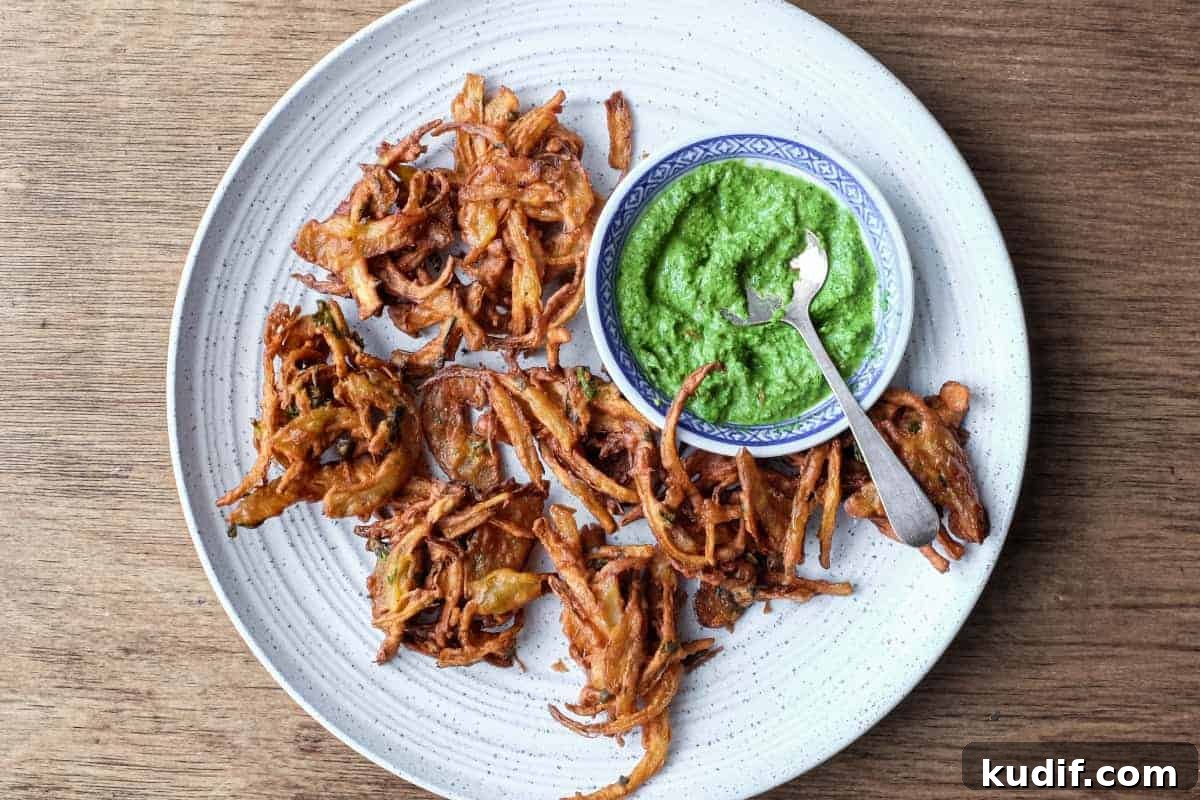 Crispy Onion Pakoda Recipe: Indian Onion Fritters Guide 12 Fried onion pakodas on a platter with a small bowl of coriander chutney and a small spoon.