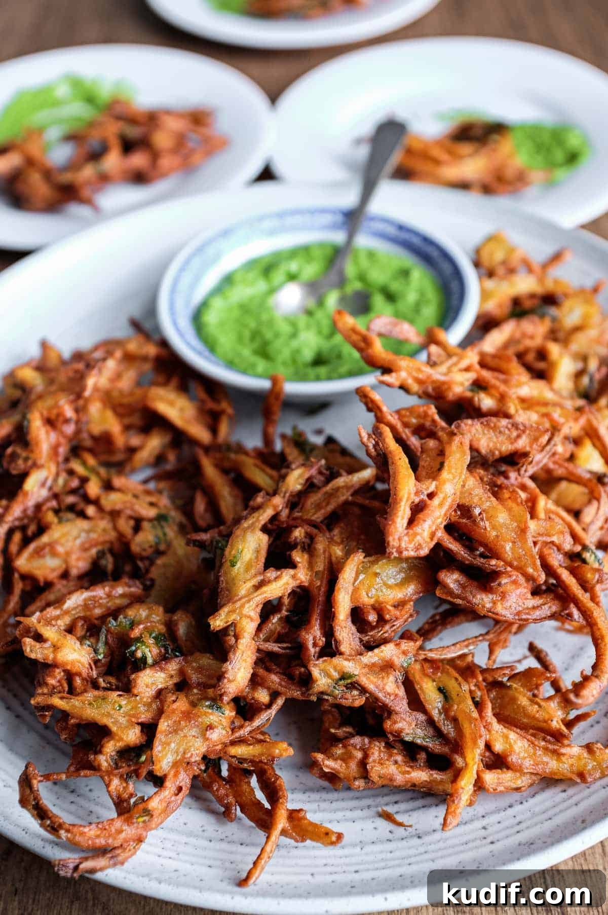 Onion Pakoda | cookeatworld.com Fried onion pakodas on a platter with a small bowl of coriander chutney and a small spoon.