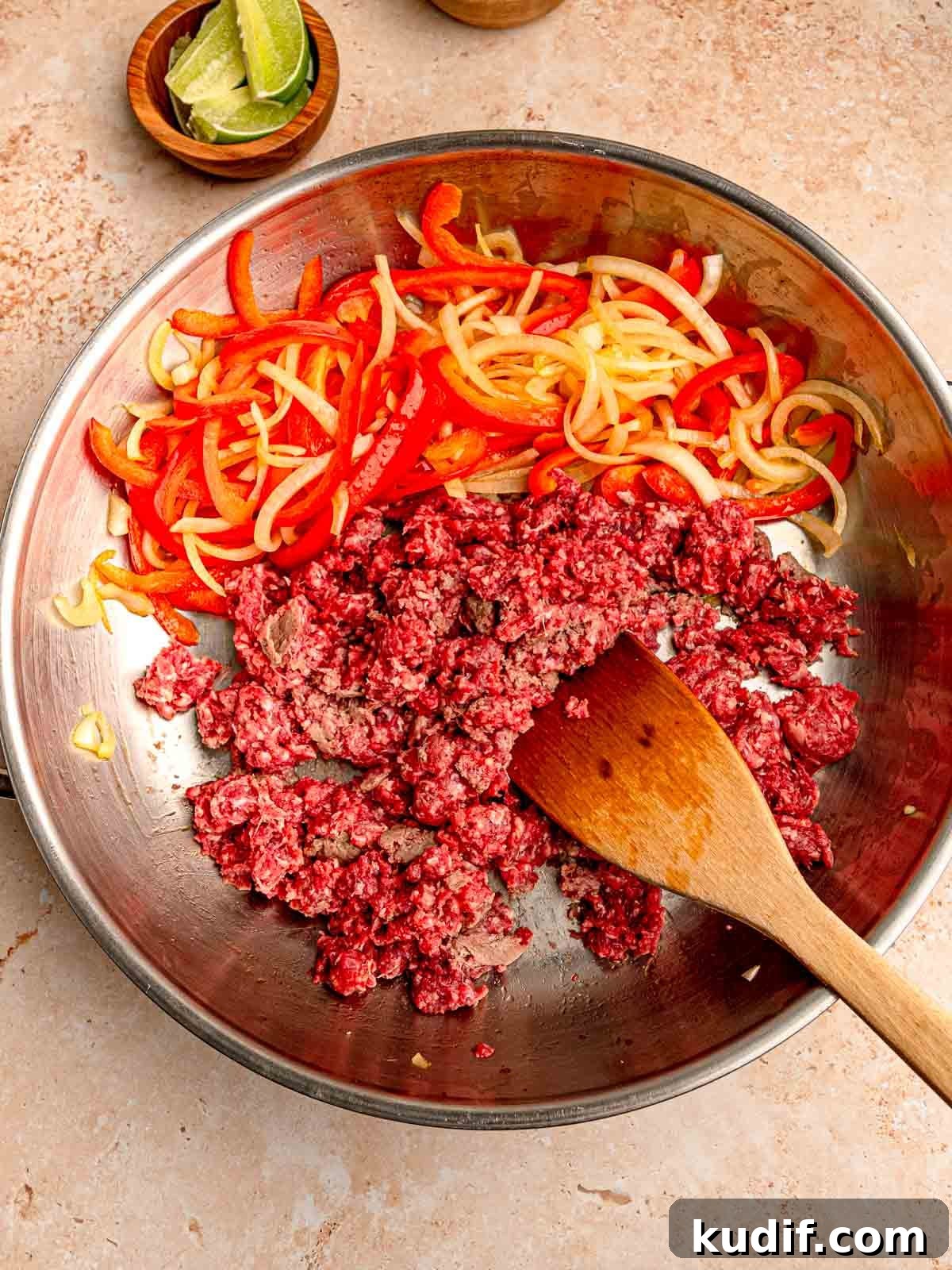 A metal pan with ground beef, sliced red bell peppers, and onions being cooked together with a wooden spatula resting on the side of the pan.