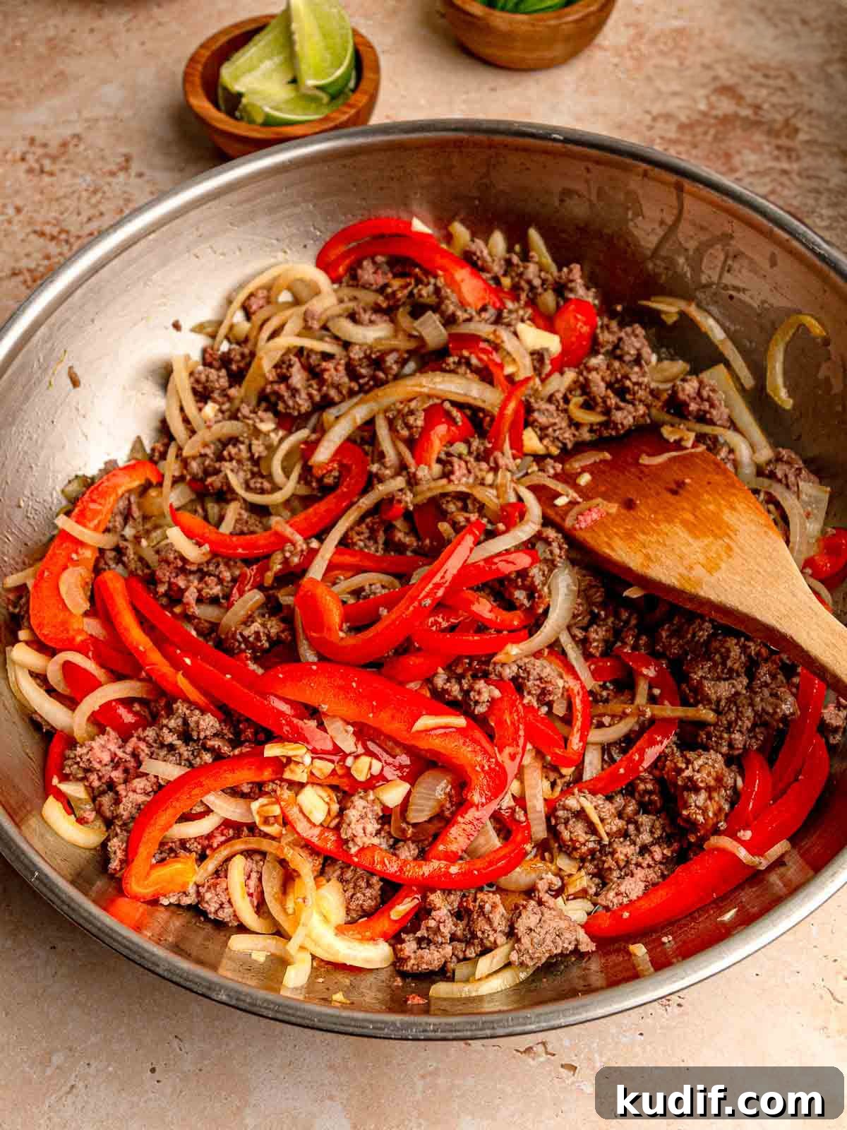 Cooked ground beef, red bell peppers, and onions in a metal pan, with a wooden spatula stirring the mixture.