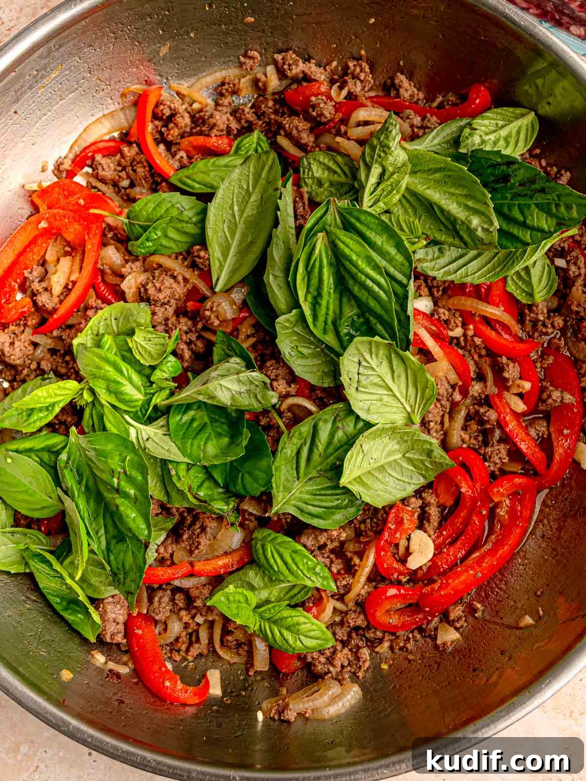 Fresh Thai basil leaves being added to a pan of cooked ground beef, red bell peppers, and onions.