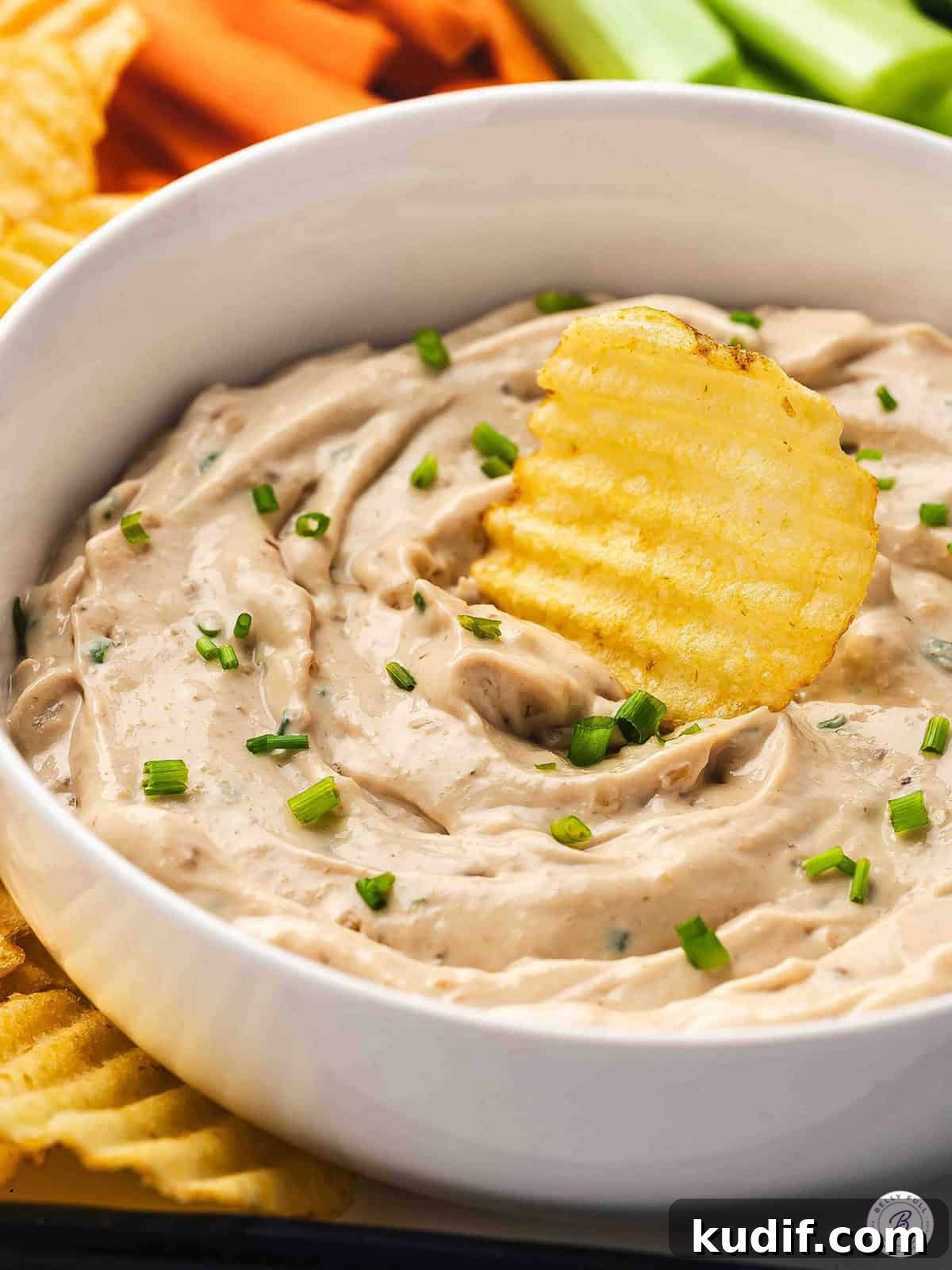 Creamy French Onion Dip Recipe for Parties and Snacks 2 French onion dip in a white bowl with chives, served with crinkle-cut potato chips and fresh vegetable sticks