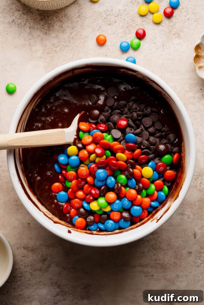 A bowl of brownie batter with m&ms and chocolate chips being stirred in.