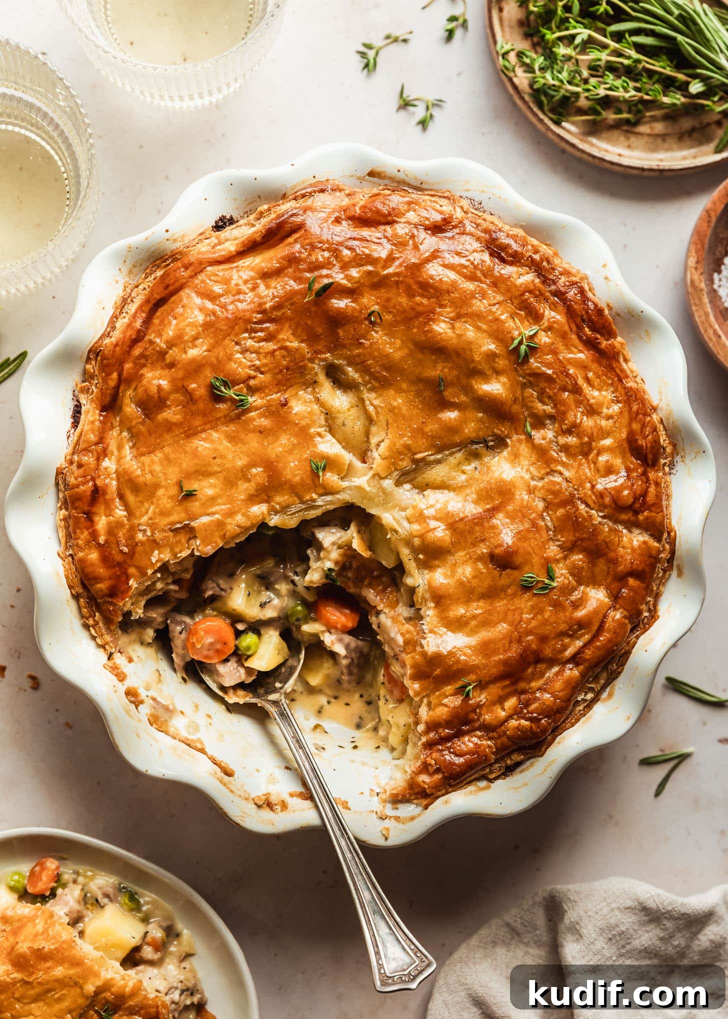Turkey pot pie with puff pastry in a white pan next to a white plate of pot pie, two glasses of white wine, beige linen, and brown plate of herbs on a beige counter.