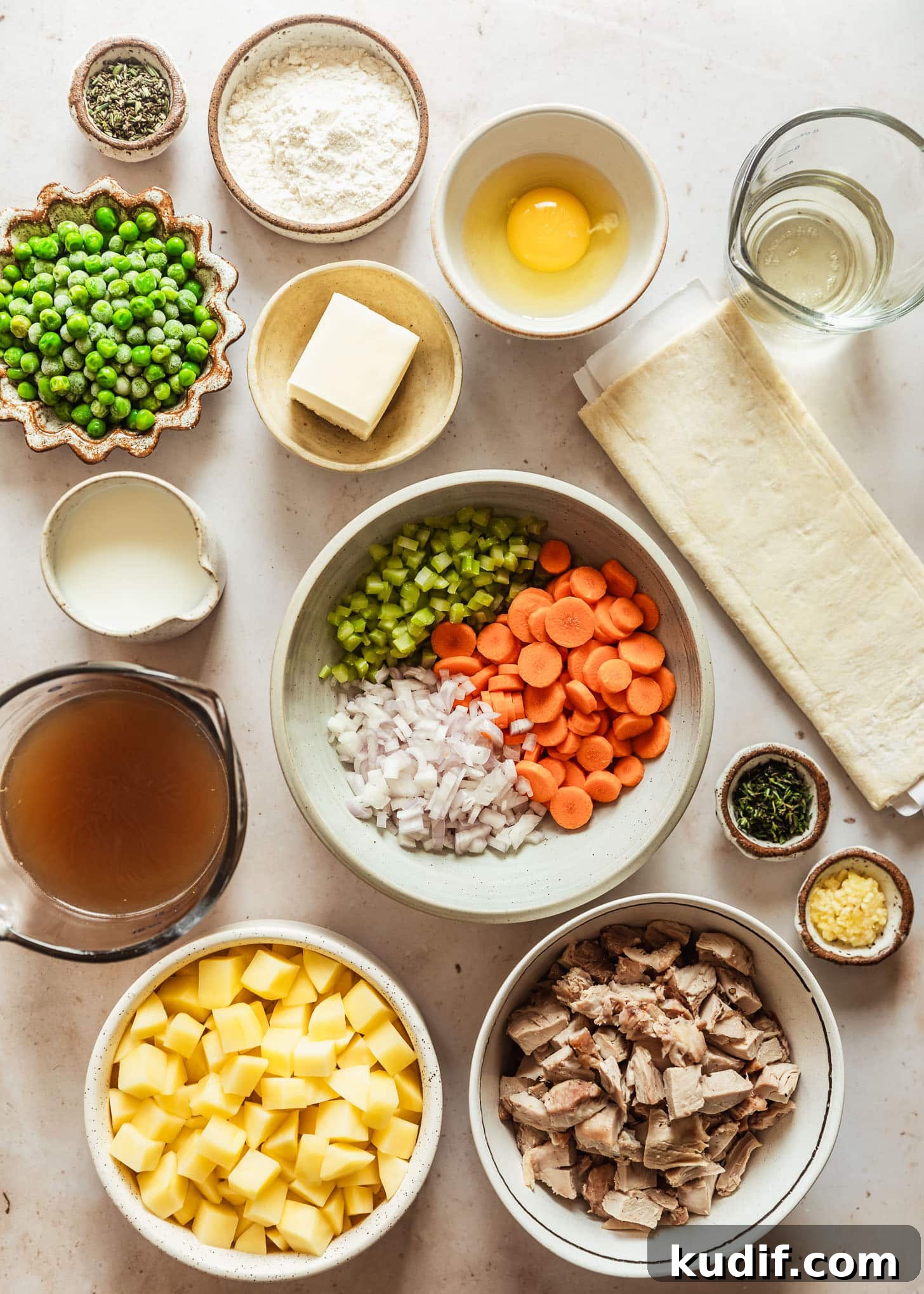 Pie dough on a beige counter next to white and brown bowls of potatoes, meat, carrots, celery, shallots, milk, herbs, garlic, peas, an egg, flour, butter, and white wine.