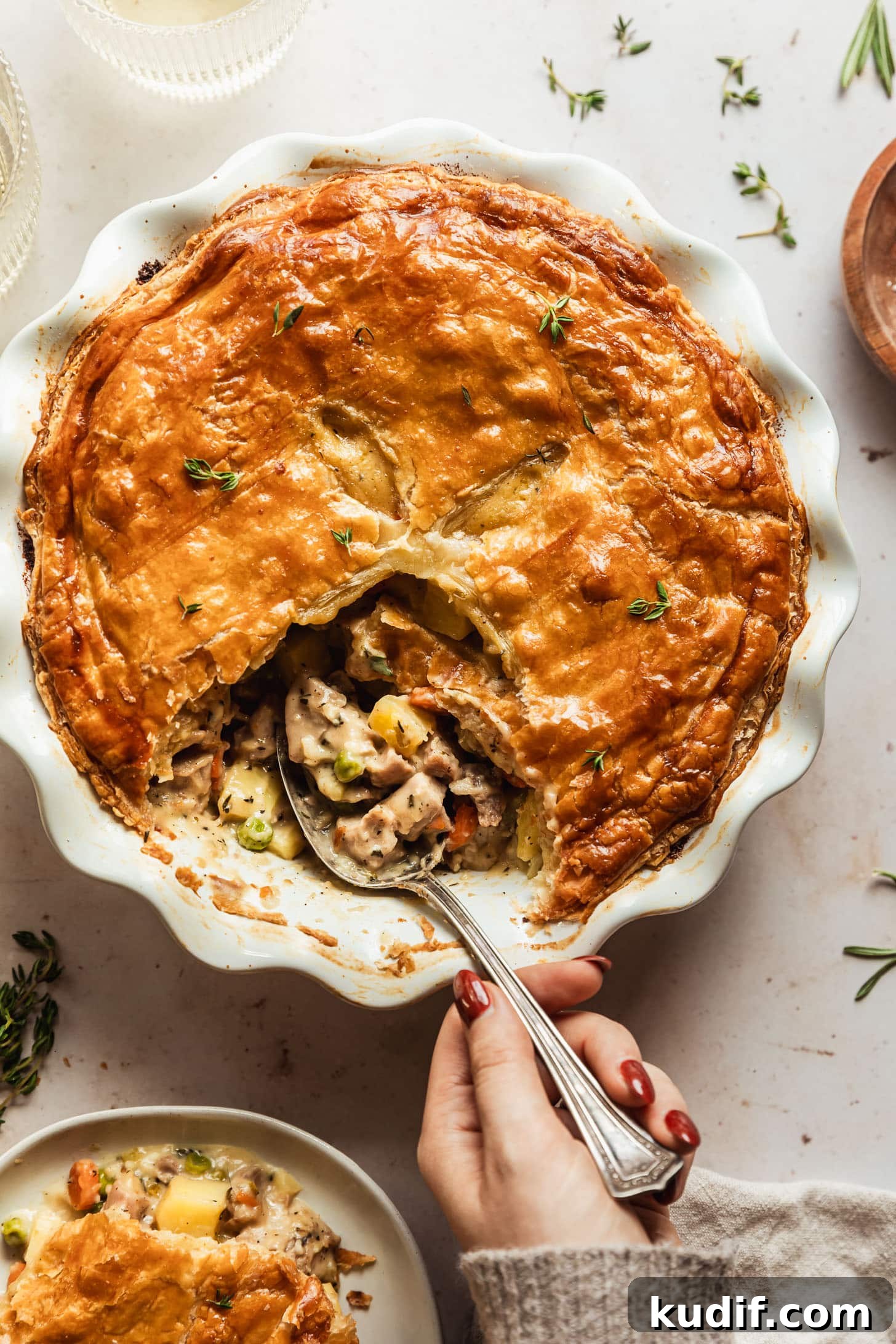 A hand grabbing a spoonful of turkey pot pie with puff pastry out of a white pan. The pan is on a beige counter next to a plate of pot pie, two glasses of white wine, and wood bowl.