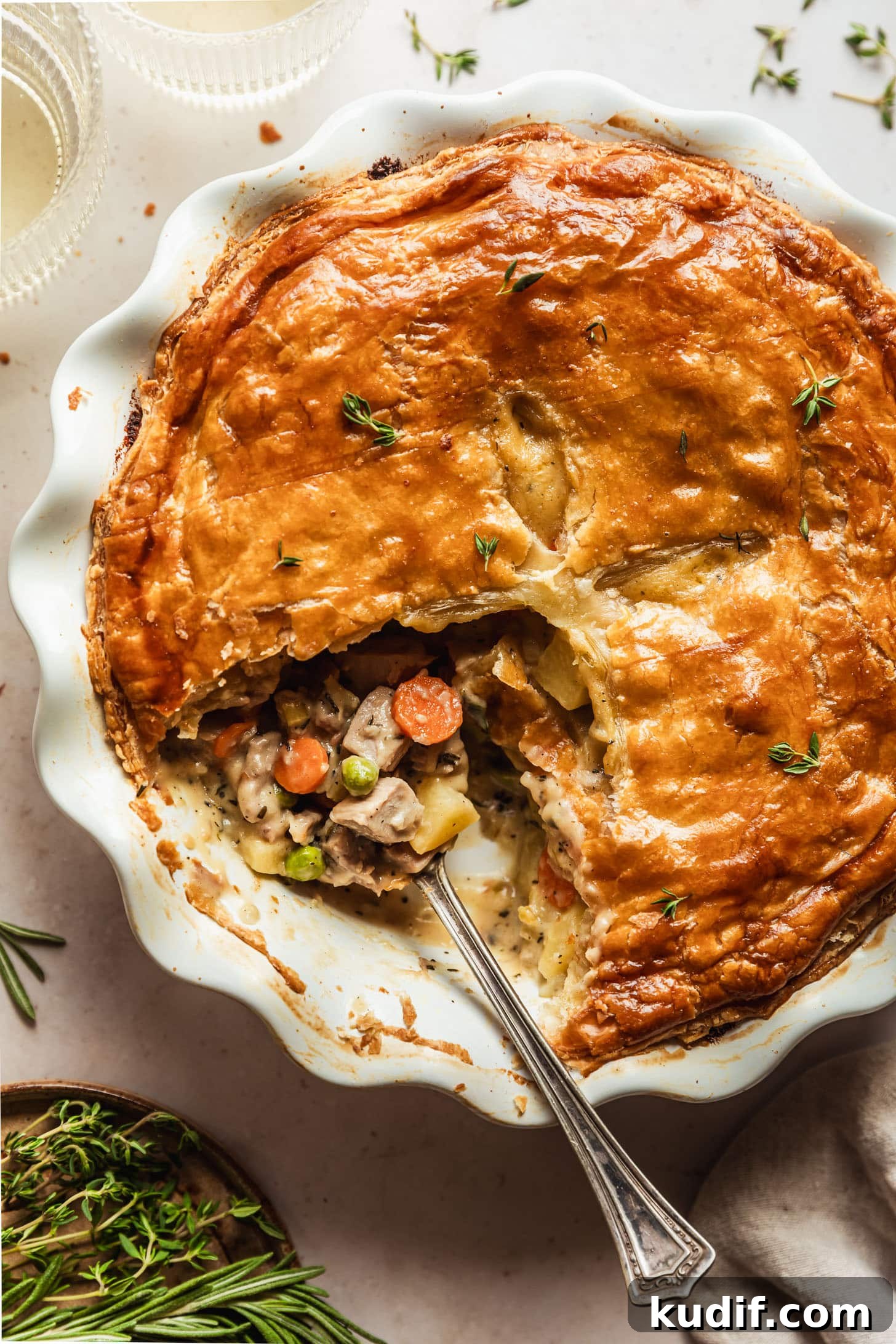 A spoon in a white pie plate of turkey pot pie with puff pastry on a beige counter next to a beige linen, glass of white wine, and brown plate of rosemary and thyme.