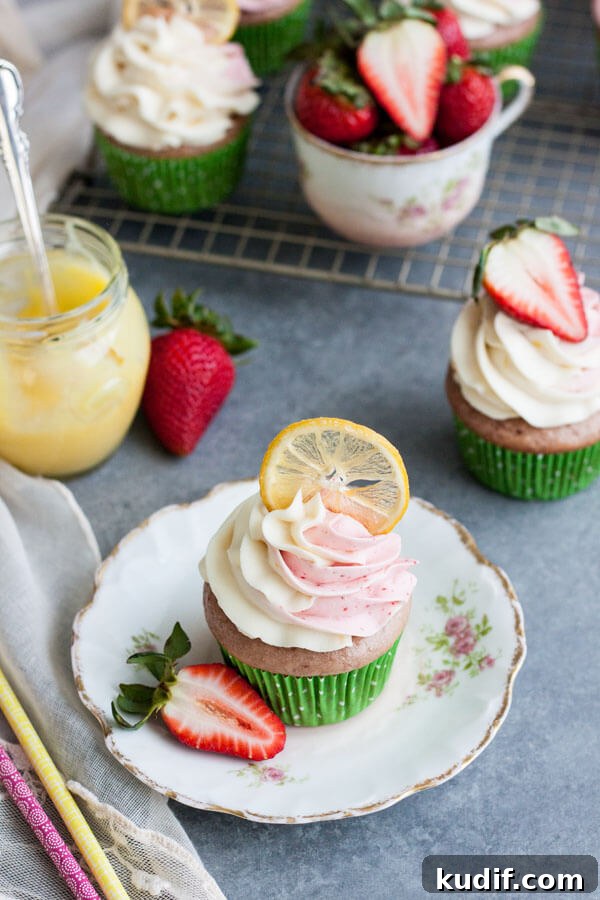 Strawberry lemonade cupcake decorated with a candied lemon slice on a plate next to a pot of lemon curd, fresh strawberries and extra cupcakes in the back