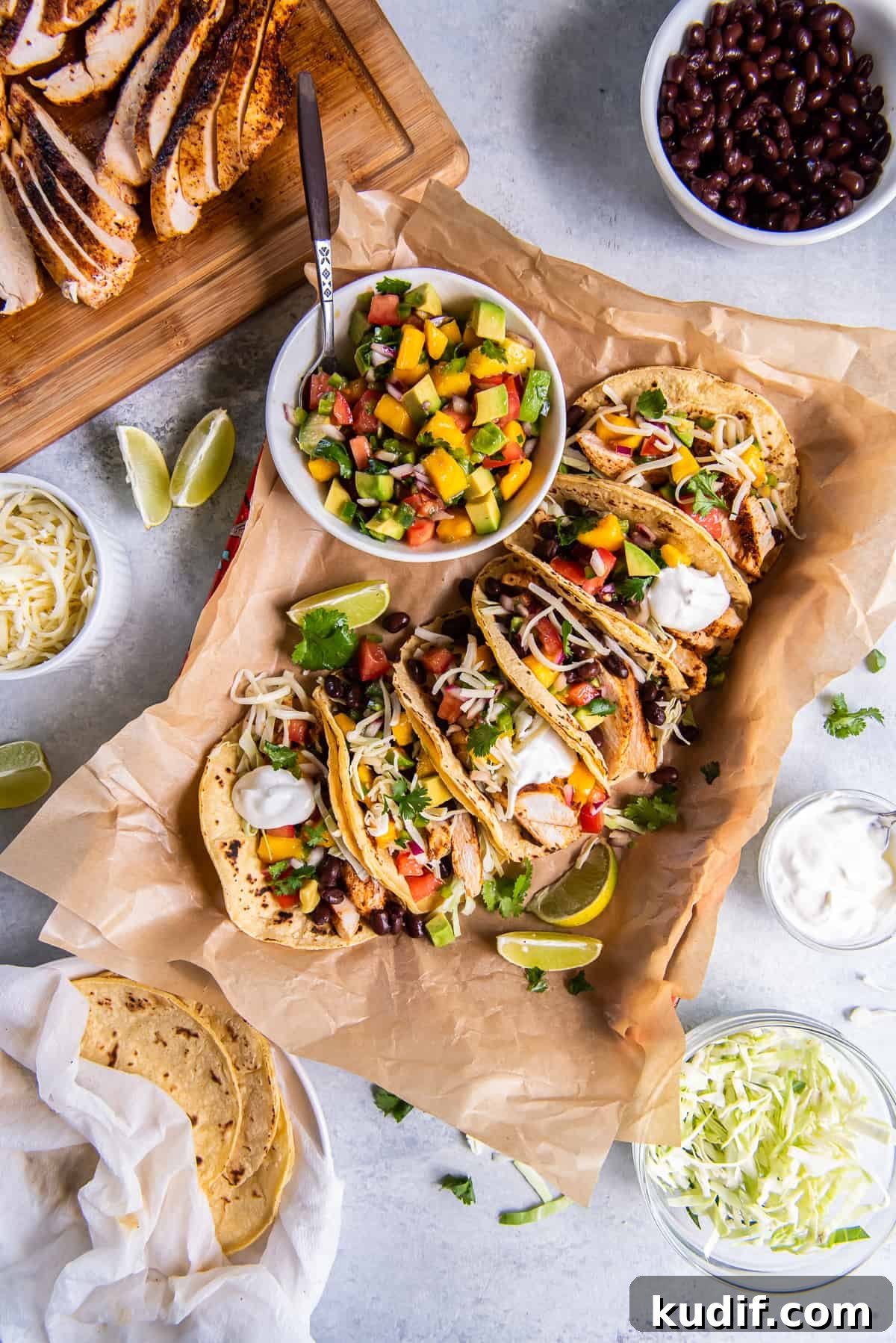 A top down shot of chicken tacos topped with mango salsa on parchment paper surrounded by bowls of ingredients.